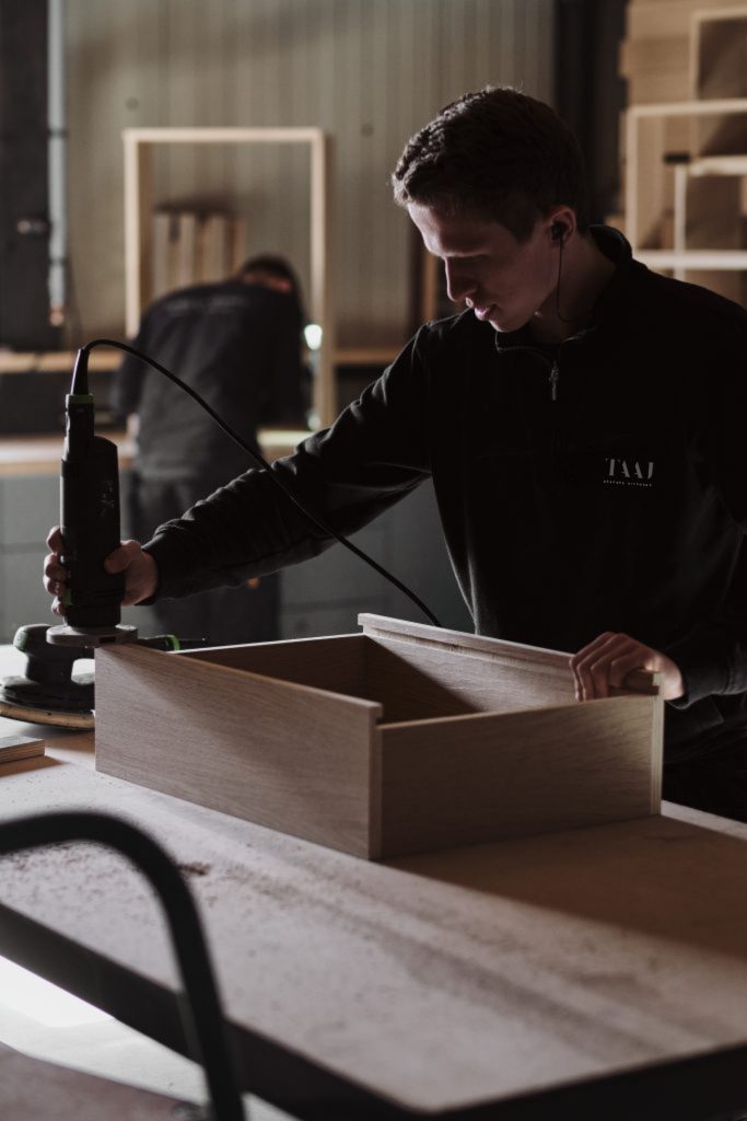 A person wearing ear protection uses a power tool to work on a wooden box in a workshop, crafting elements for bespoke luxury kitchens while another individual is also busy.