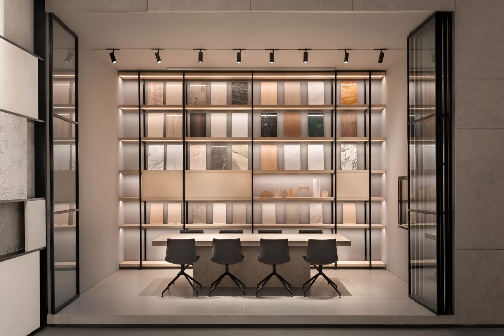 A modern showroom with a backlit display of various stone and wood samples, showcasing luxury bespoke kitchens in the UK, and a long table with four chairs in front of it.