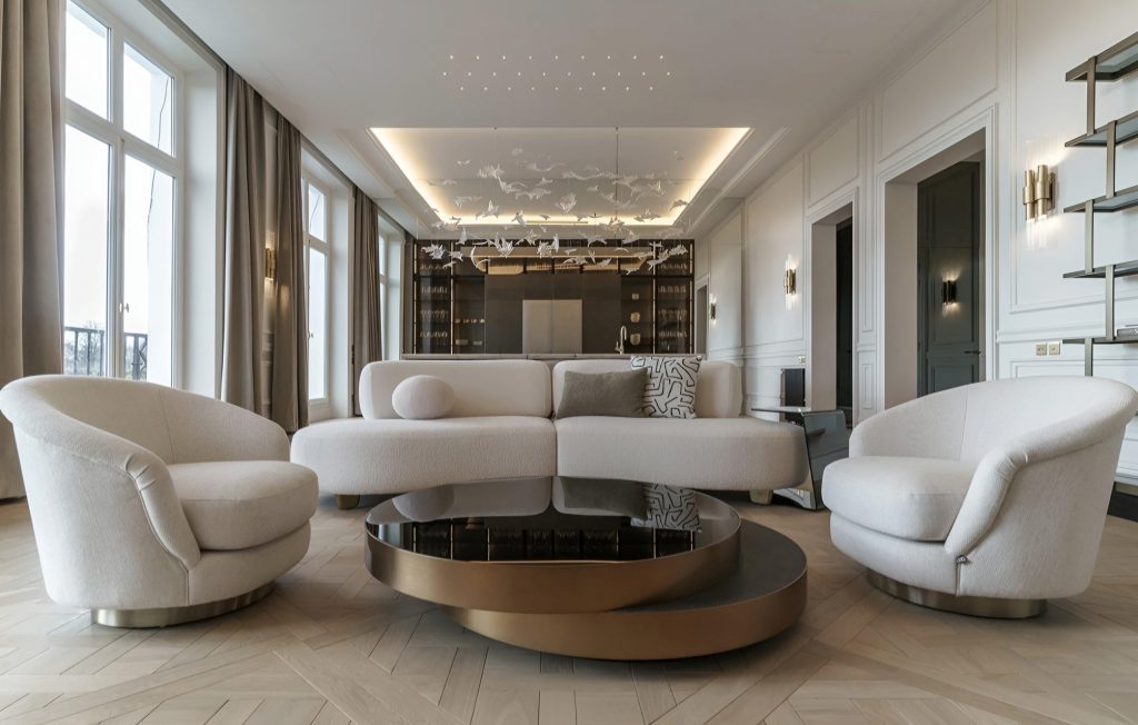 Modern living room with white furniture, including a sectional sofa and two round chairs. A round black and gold coffee table is in the center, complemented by floor-to-ceiling windows and a sleek design that perfectly pairs with an adjacent luxury kitchen renovation.