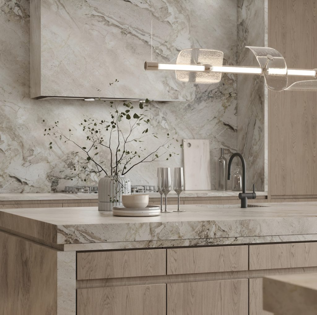 Experience a luxury kitchen with light marble countertops and backsplash. Wooden cabinets, vases with a plant, and glasses adorn the countertop. A contemporary light fixture illuminates this bespoke luxury kitchen.
