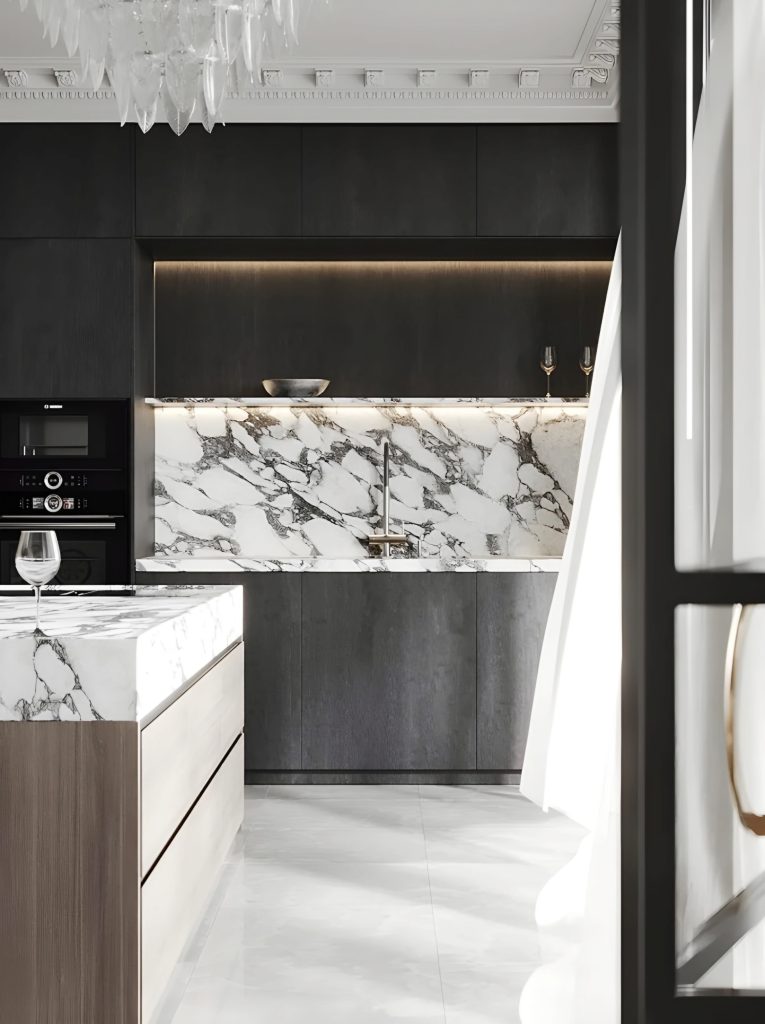 This modern kitchen boasts dark cabinetry, marble countertops, and a matching marble backsplash. A glass of wine sits on the countertop, while an elegant chandelier hangs above, showcasing the luxury and attention to detail that defines bespoke luxury kitchens.