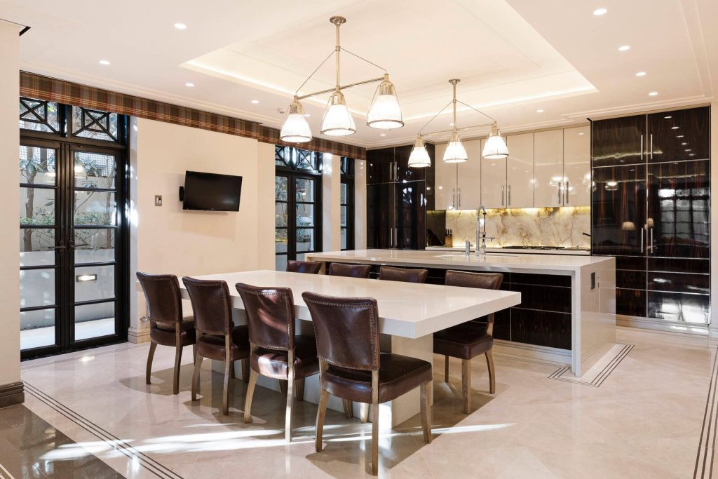 A modern kitchen features a large island with six brown leather chairs, stainless steel appliances, marble flooring, pendant lights, and floor-to-ceiling windows—an epitome of bespoke luxury kitchens crafted by a top kitchen renovation company.