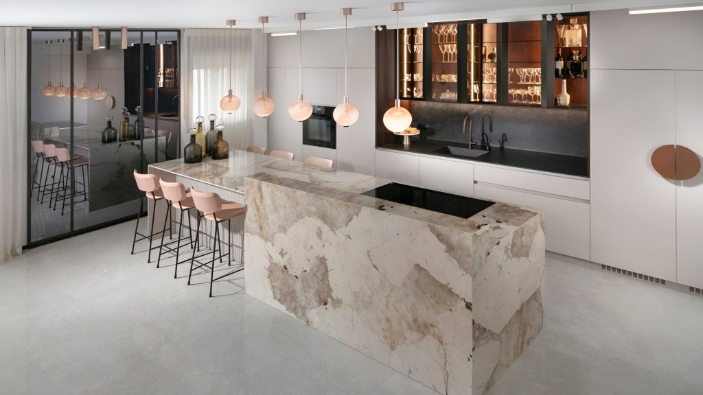 A luxury kitchen renovation featuring white cabinets, a marble island with integrated stovetop, pink bar stools, pendant lights, and a mirrored wall.