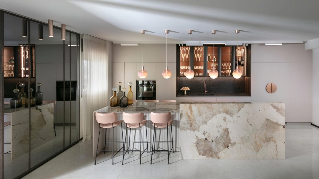 A modern kitchen featuring a marble island, four pink bar stools, hanging pendant lights, glass cabinetry, and a mirrored wall. Perfect for those seeking the expertise of a luxury kitchen renovation company.