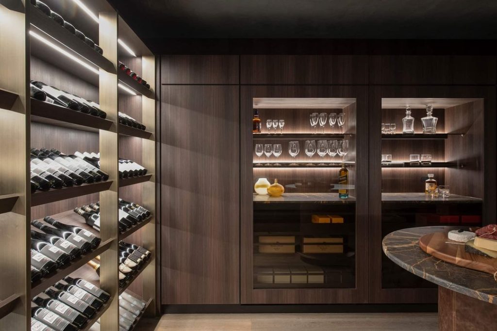 A modern wine cellar with illuminated wooden shelves holding wine bottles, a glass cabinet with various glasses and decanters, and a round table with charcuterie items complements the luxury kitchen renovation.