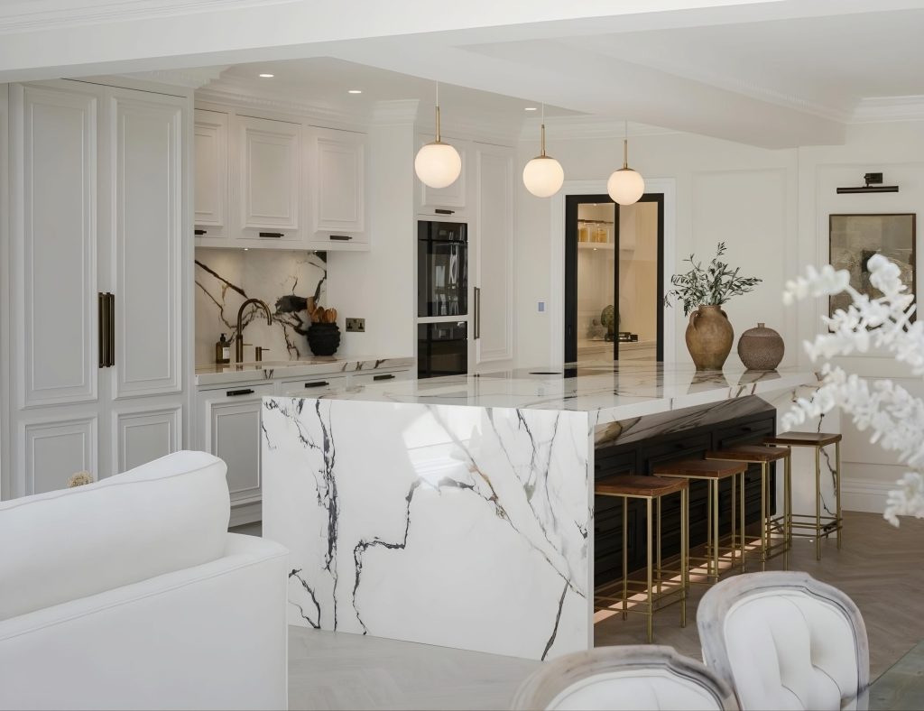 Modern kitchen with white marble countertops and island, wooden barstools, white cabinetry, pendant lights, and decorative vases. A luxury kitchen renovation showcasing a white sofa and chairs partially visible in the foreground.