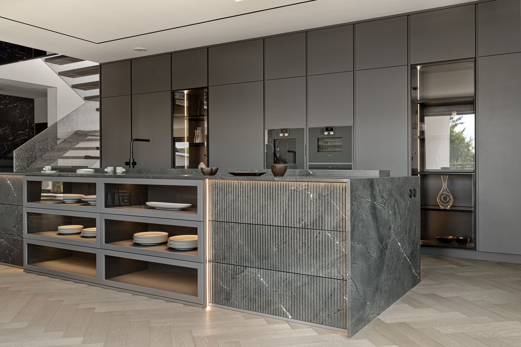 Modern kitchen with sleek, dark cabinets, an island with open shelving displaying dishware, built-in ovens, and a minimalist design. The decor features gray tones and wood-pattern flooring.