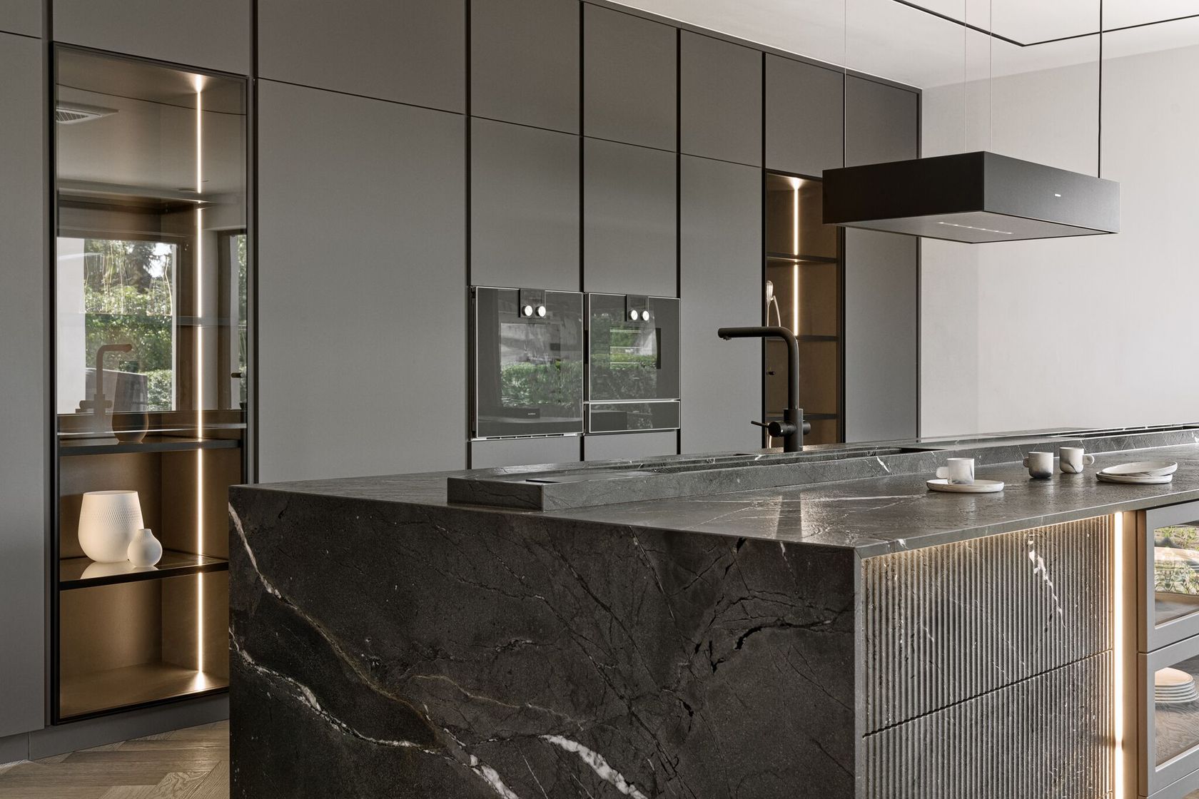 Modern kitchen with dark marble island, integrated appliances, and sleek gray cabinets. Minimalist design with ambient lighting and decorative objects on display shelves.