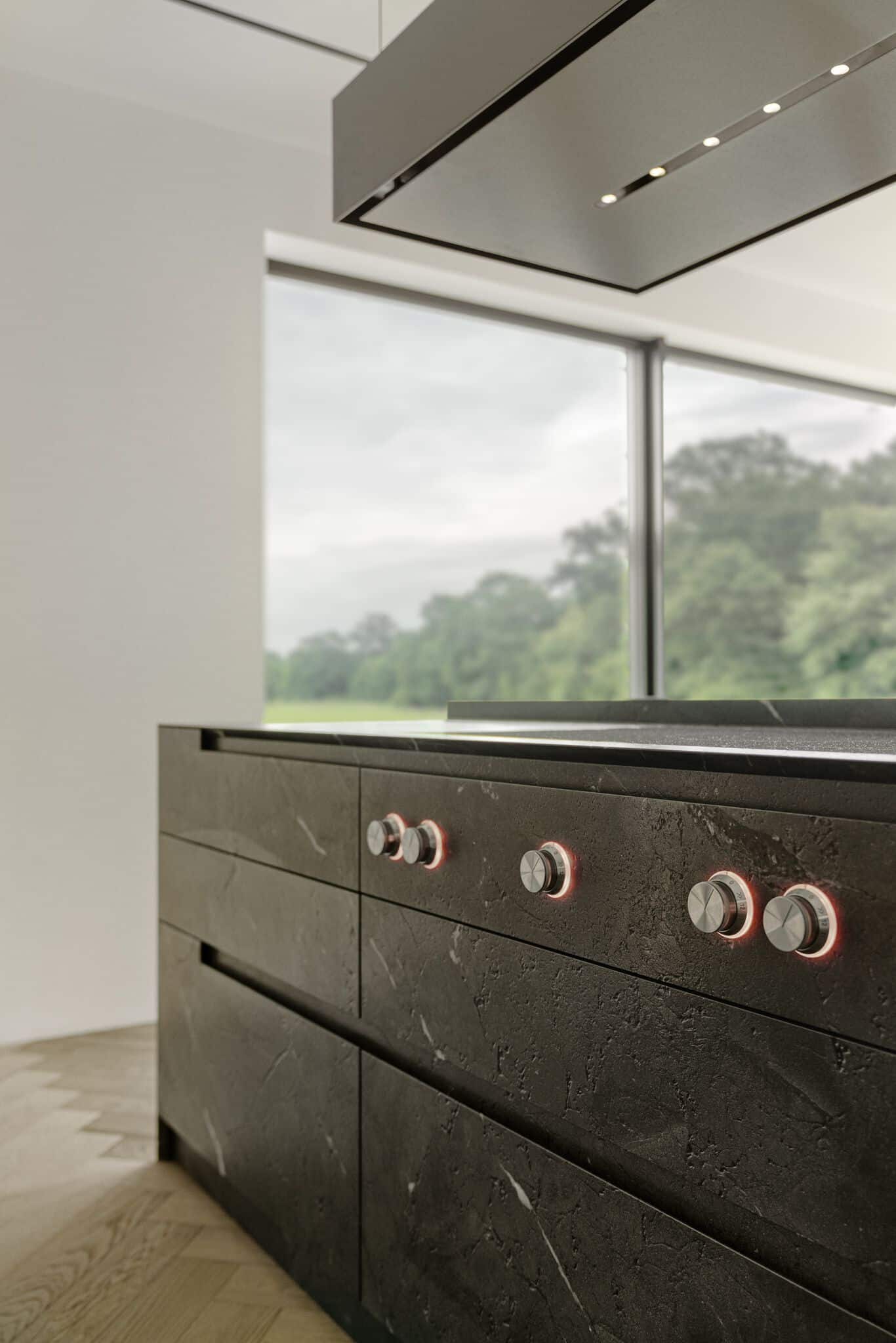 Modern kitchen with sleek dark cabinetry featuring metallic knobs, set against a window showcasing a view of greenery. Light wood flooring complements the minimalist design.