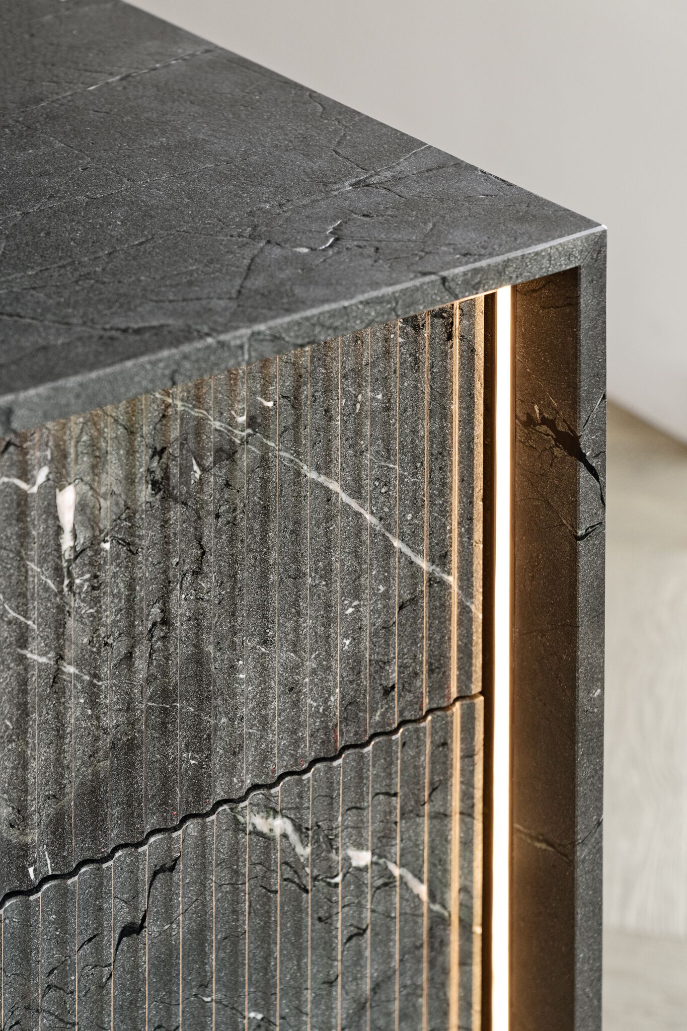 Close-up of a textured dark gray stone surface with vertical grooves. A warm, vertical LED light strip is embedded along the corner edge, enhancing the stone's texture and color.